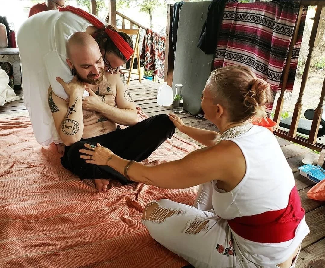  Shea Bilé sits on a pink blanket on the floor, smiling as a woman with hair in a bun and wearing a white tank top and ripped pants reaches out to hold his leg. A woman with a red headband leans over him from behind. 