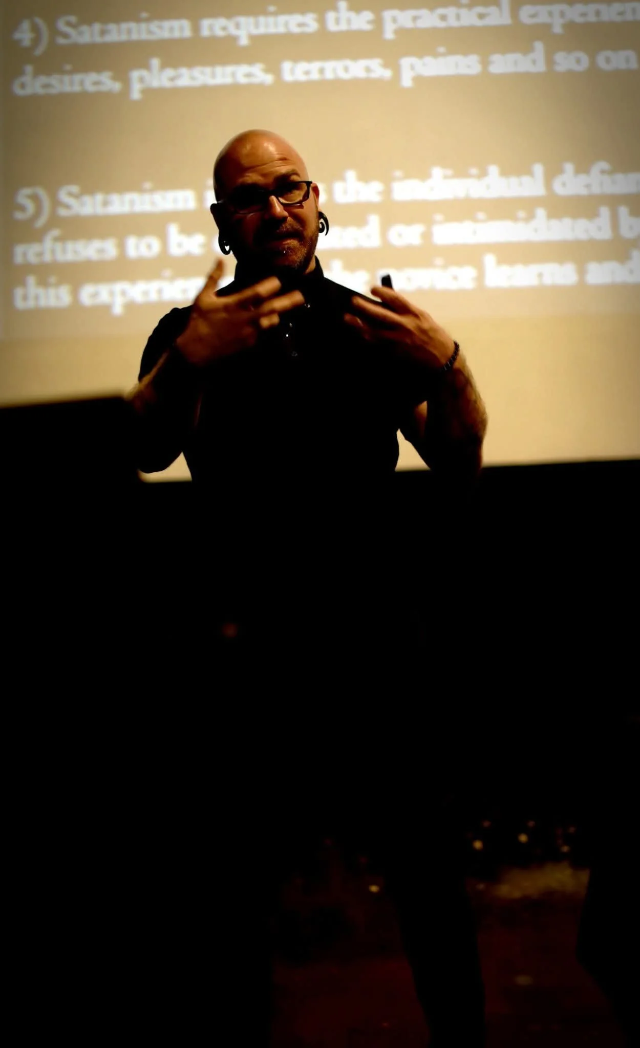 Shea Bilé is standing in front of a projected presentation, gesturing with his hands during a lecture or presentation.