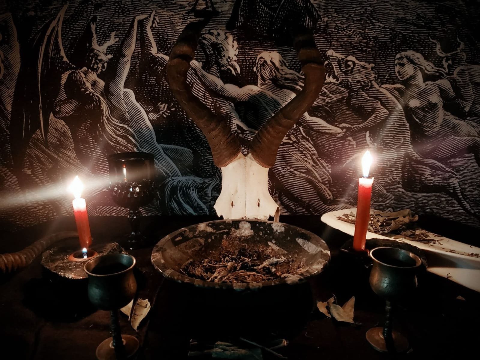 A ritual setup with a skull of a ram with large curved horns, two lit red candles, metal goblets, a large bowl with ashes or earth, and a decorative cloth with an image of the Witch's Sabbath.
