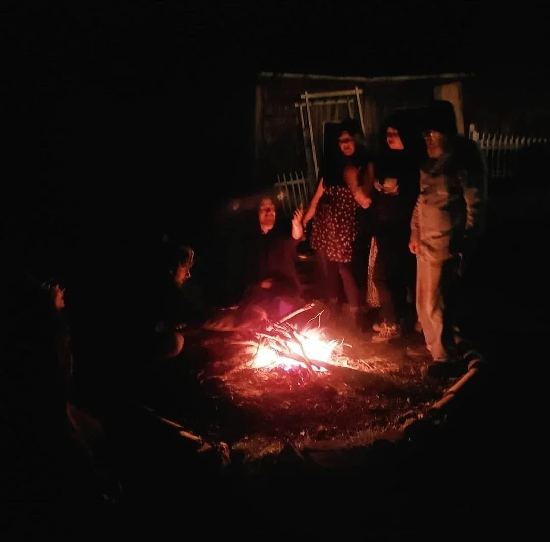 Shea Bilé and a group of people are gathered around a campfire at night, standing and sitting nearby, with a wooden fence and structure in the background.