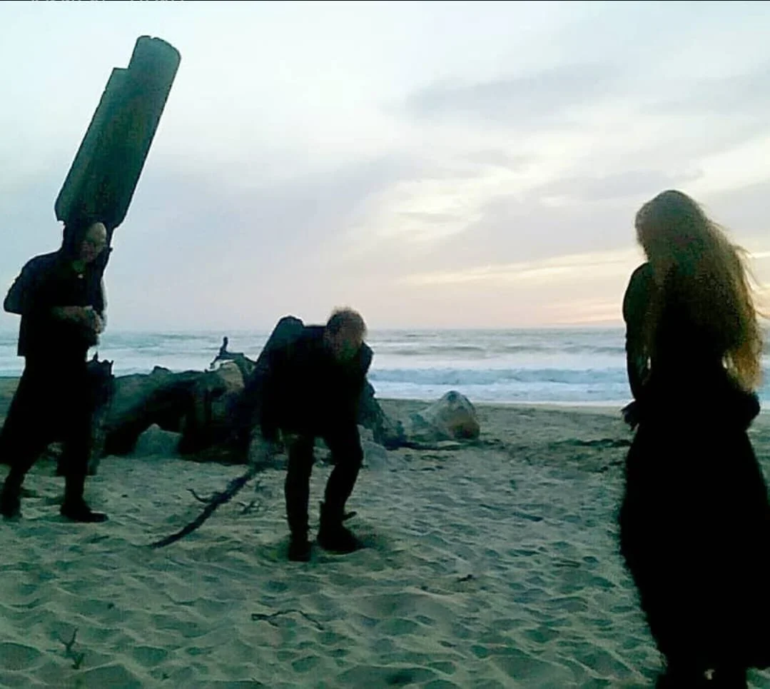 Shea Bilé and two other people on a beach during sunset, one is crouched holding a rope, and another is standing with long hair. The ocean is visible in the background.