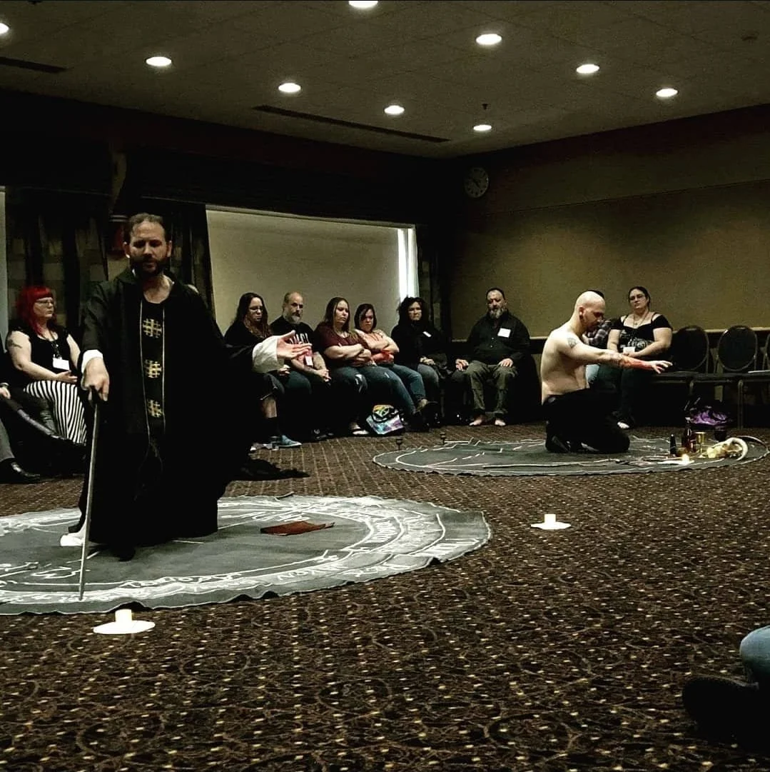 A group of people sitting in a circle in a dimly lit room watching Shea Bilé and Jack Grayle on a circular mat, Shea is kneeling and Jack is standing with a sword, with drinks and objects on the floor.