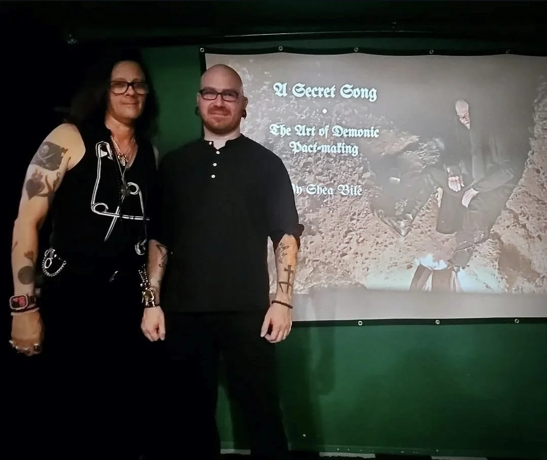Shea Bilé and Sal Santoro stand in front of a screen displaying the text 'A Secret Song, The Art of Demonic Pact-Making, by Shea Bile.' They are posed indoors with a green backdrop.