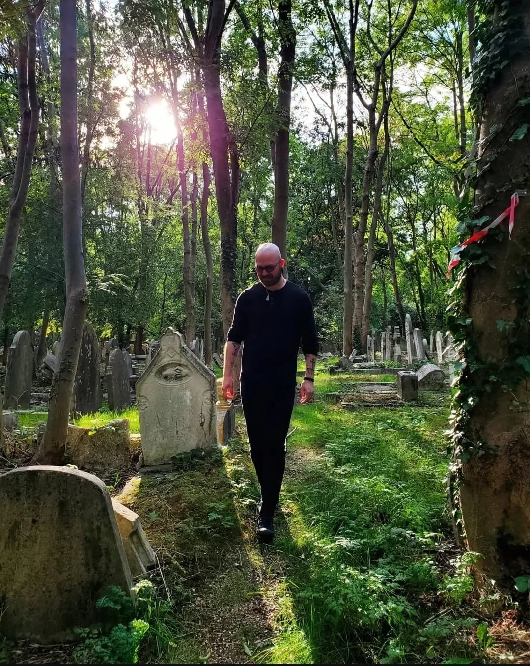 Shea Bilé walking on a narrow path in a wooded graveyard with old tombstones and sunlight filtering through the trees. Paris, France. 