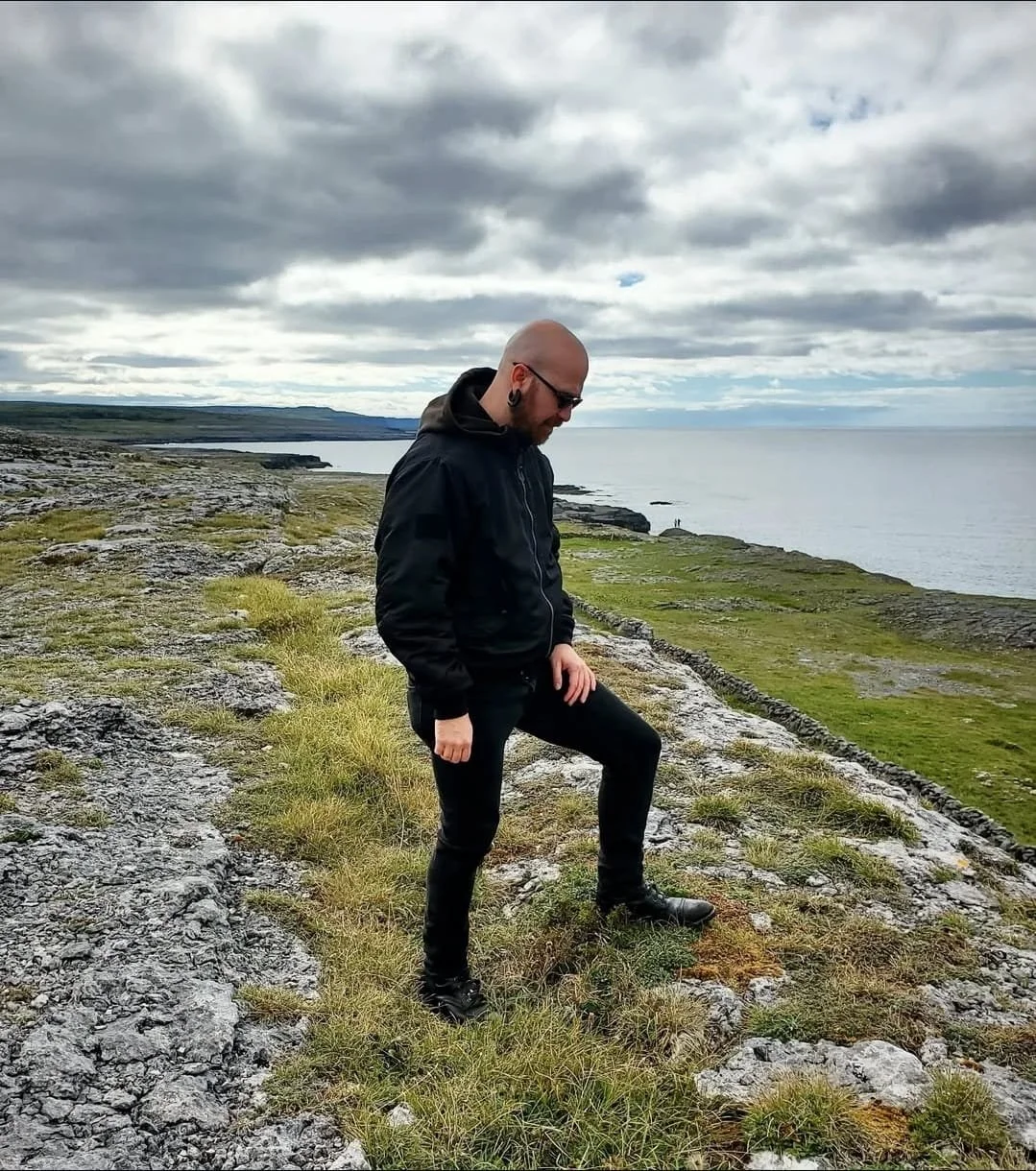 Shea Bilé standing outdoors on rocky terrain with grassy patches, overlooking the ocean under cloudy skies. Ireland.