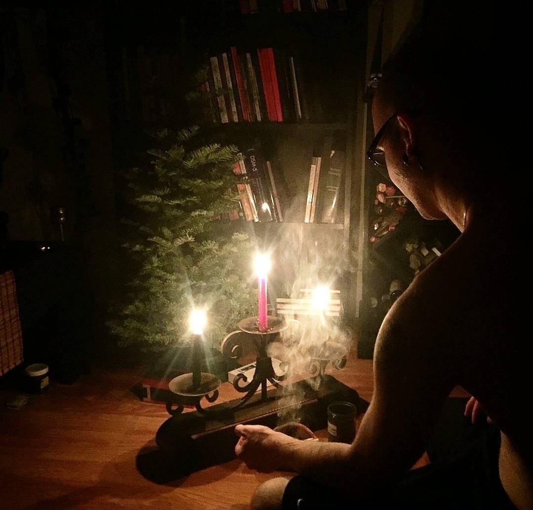 Shea Bilé sits in a dimly lit room with a lit candle on a candelabrum. There is a bookshelf with books and a Christmas tree in the background, and smoke is rising from the candle.