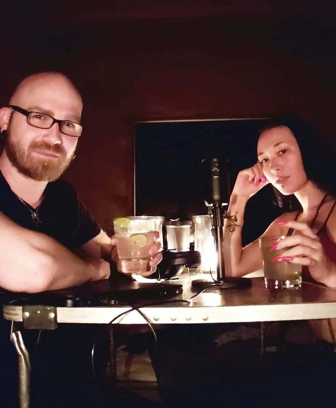Shea Bilé and Luzia Lowe sit at a table with drinks, in a dimly lit room with a microphone between them. Recording an episode of Deferred Gnosis. California. 
