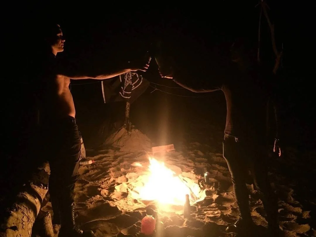 Two people standing near a campfire at night on the beach, with sand and some logs visible.
