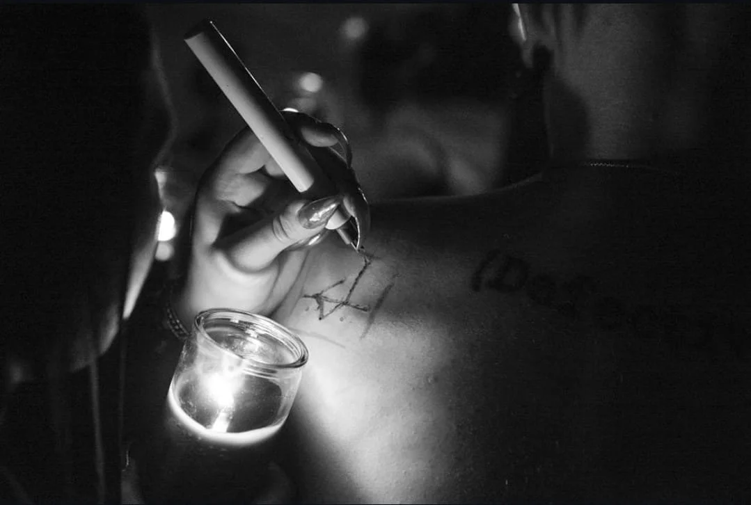 Luzia Lowe uses an electric cauterizing device to burn a sigil into Shea Bilé's back, illuminated by a small candle in a glass jar.