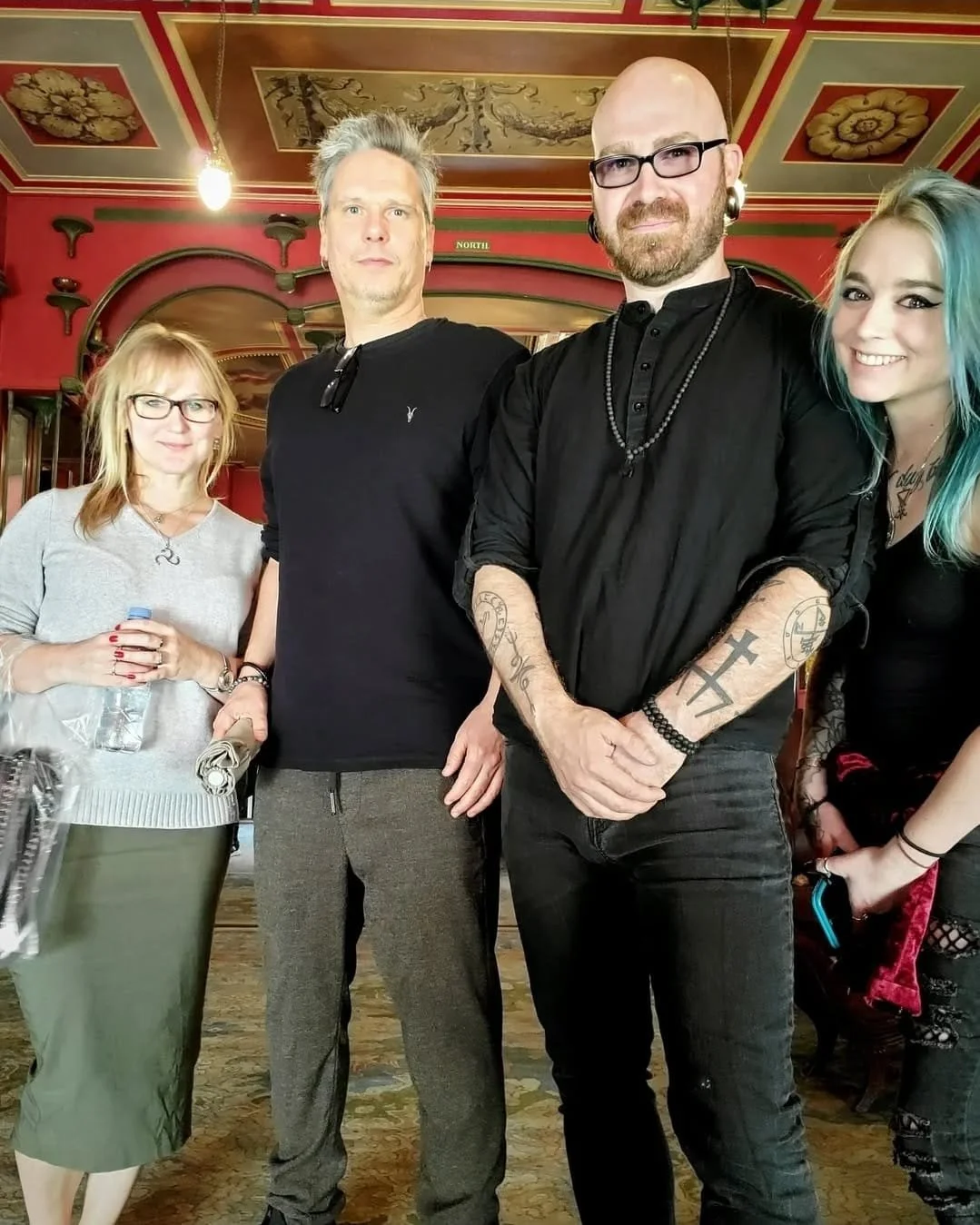 Shea Bilé and David Beth standing together in a decorated room with a painted ceiling and red walls. The group includes a woman with blonde hair, and a woman with colorful blue-green hair. London, England.