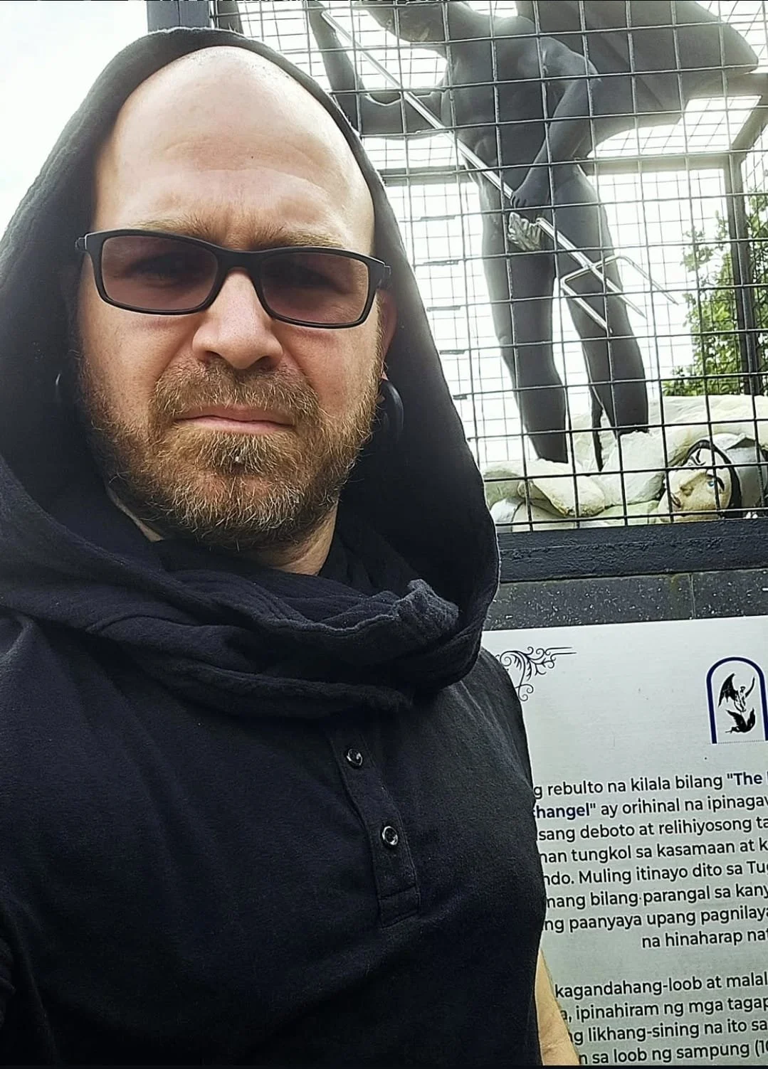 Shea Bilé wearing a black hoodie, taking a selfie in front of a cage with a statue of Satan defeating the archangel Michale. A plaque has text in Tagalog. Manila, Philippines.