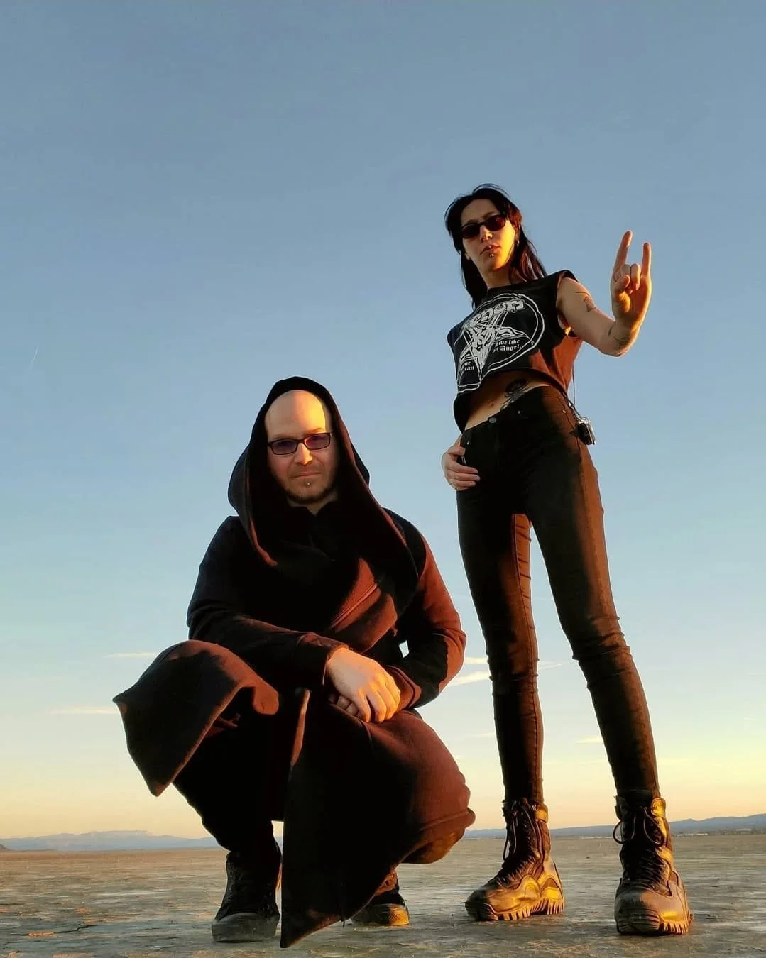 Shea Bilé and Luzia Lowe standing outdoors on a flat surface during sunset. Both are wearing dark clothing, including boots, with Luzia showing the sign of the horns. The sky is clear with a few clouds on the horizon. Mojave Desert, California.