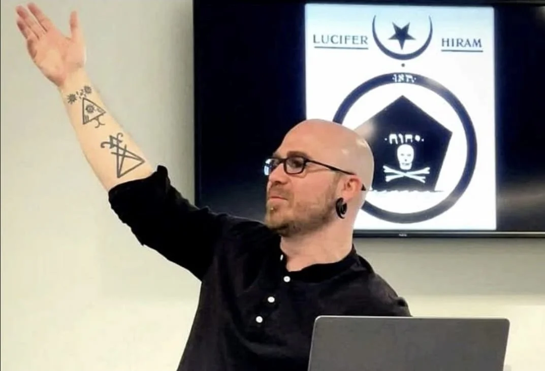 Shea Bilé raises one arm, a screen behind him displays an occult text. Lucifer Hiram.
