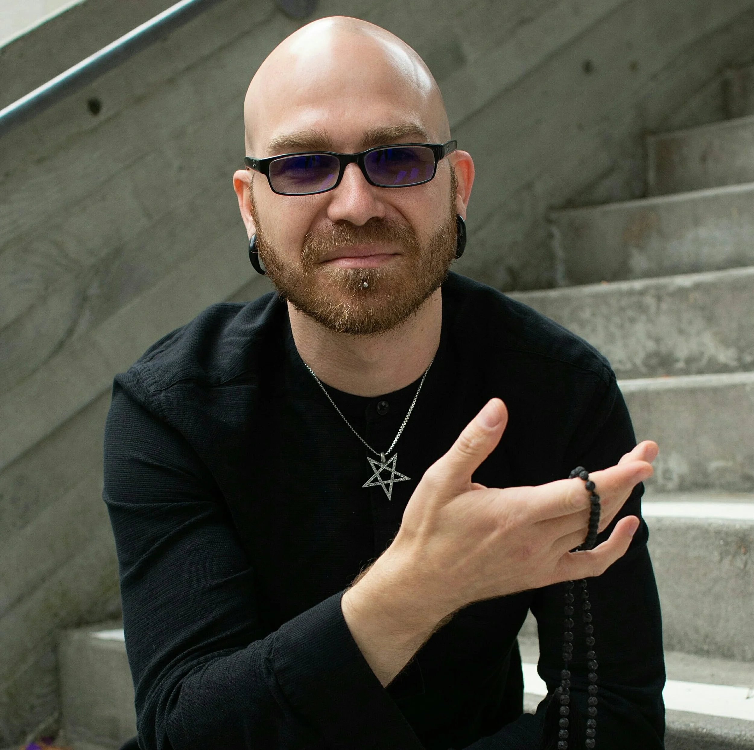 Author Shea Bilé (Shea Bile) seated on concrete stairs in a portrait photo, wearing an inverted pentagram necklace and holding a black lava stone mala.