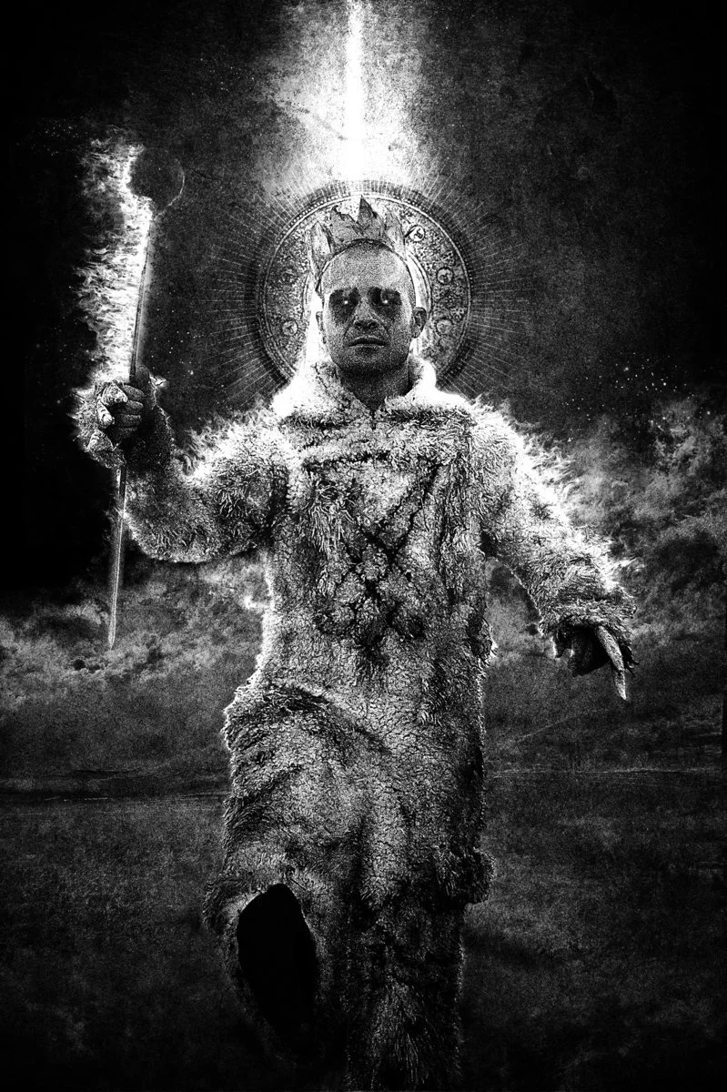 Ceremonial portrait of Shea Bilé (Shea Bile) wearing a Max suit inspired by Where the Wild Things Are, holding a staff, with a Lucifer sigil burned into the front of the garment and a radiant halo-like motif behind him in a dark ritual setting.
