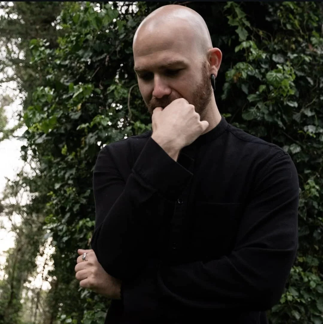Shea Bilé in a black shirt standing outdoors near greenery, with one hand touching his chin in a thoughtful pose. San Francisco, CA.