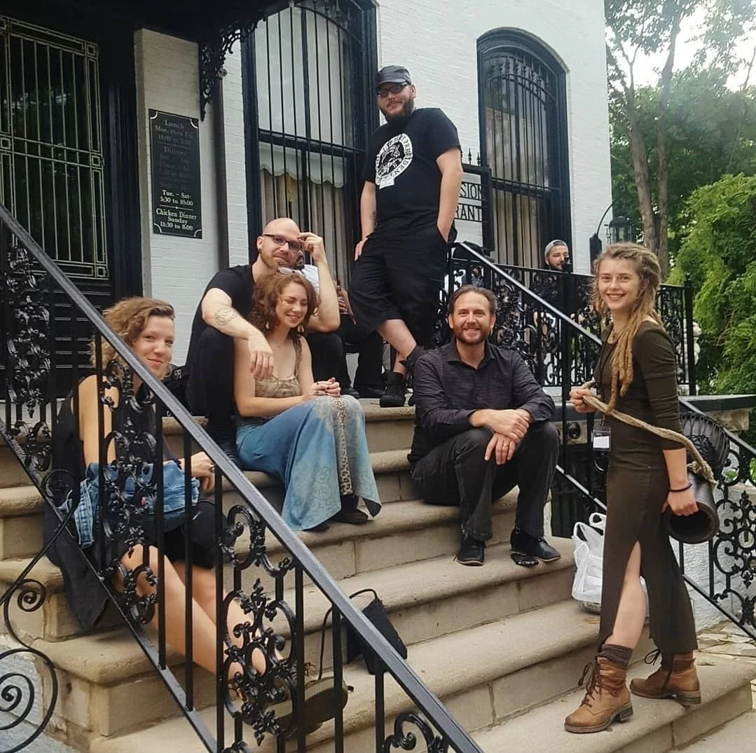 Shea Bilé and seven other people, six sitting on steps and one standing, pose on the steps of a building with black wrought iron railings. The group is casually dressed, smiling, and appears to be socializing. Lemp Mansion. St. Louis, MO.