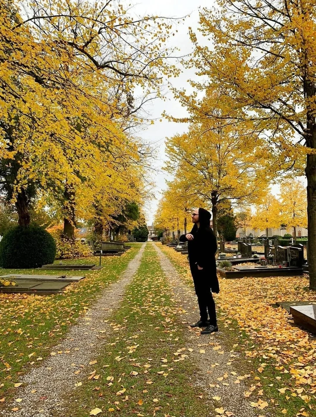 Shea Bilé is wearing black clothes and a hoodie standing on a gravel path in a cemetery with yellow autumn trees and gravestones on both sides. Leuven, Belgium.