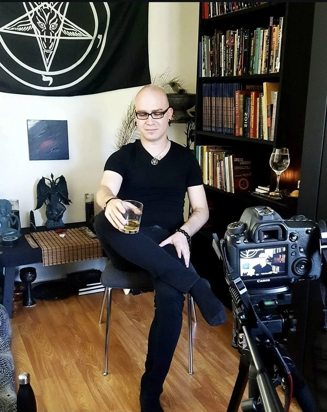 Shea Bilé sits on a stool in a room, holding a glass of whiskey, with a camera recording him, against a background of bookshelves, and an altar with a Baphomet flag.