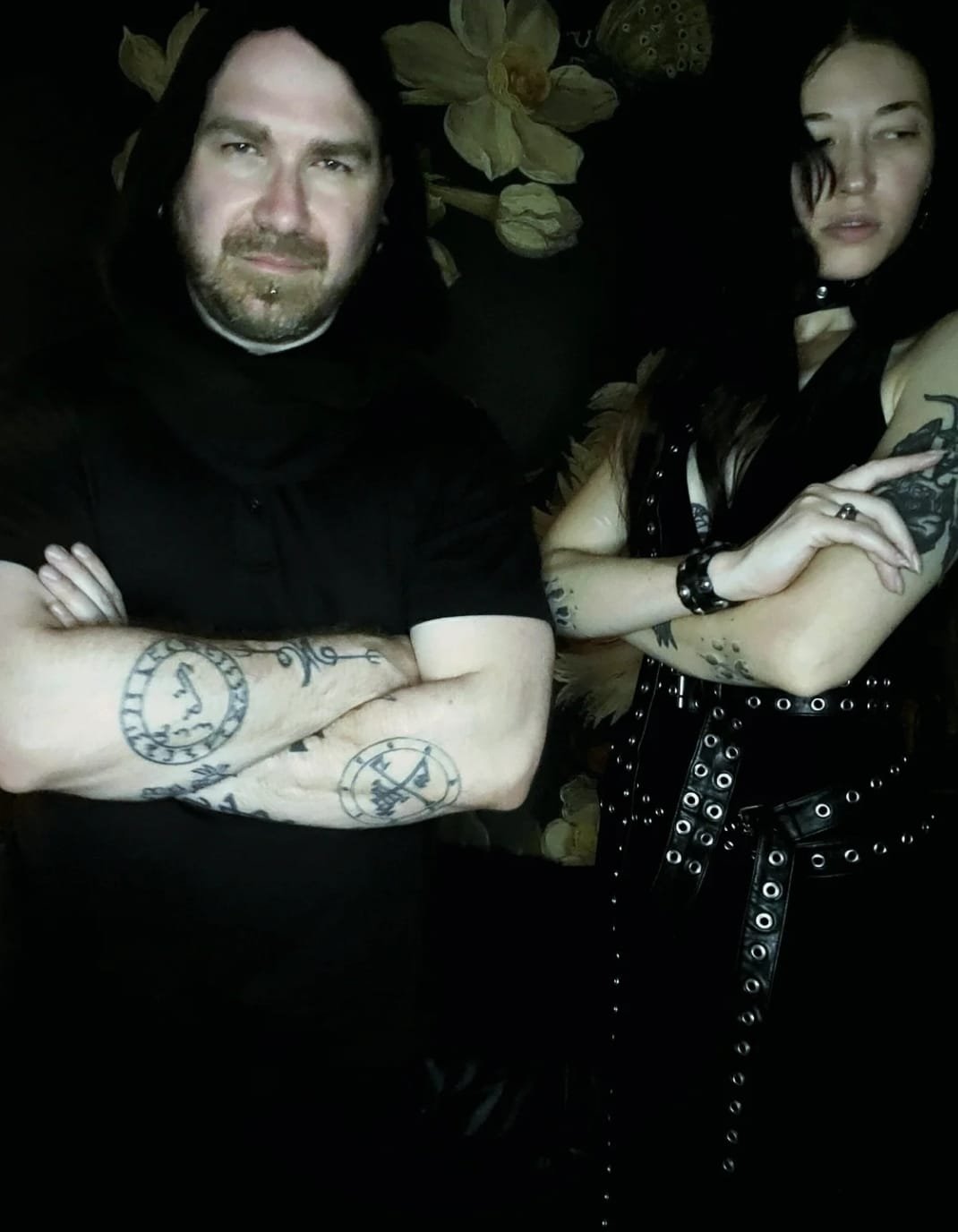 Shea Bilé standing with crossed arms. Next to him is Luzia Lowe wearing a black leather outfit with metal grommets, standing with her arms crossed. They are in front of a black background with large floral designs. Los Angeles, CA.