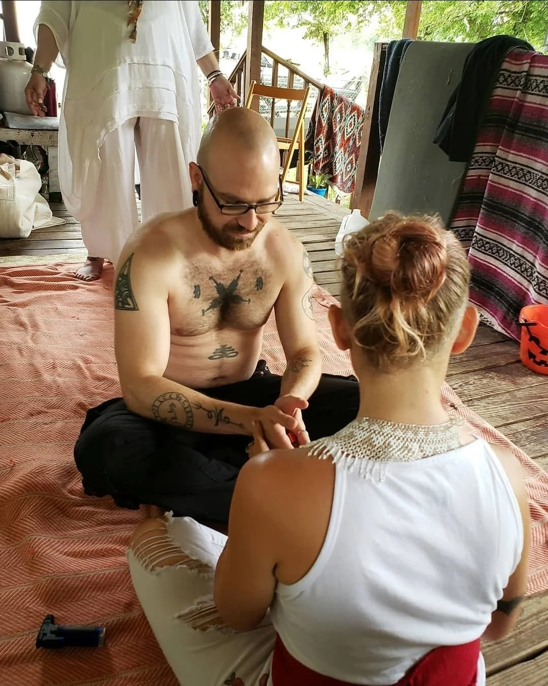 Shea Bilé sits cross-legged on a blanket, holding hands with a woman with curly hair tied up, sitting with her back to the camera. They are outdoors on a wooden porch, with a woman standing behind them, and colorful blankets hanging on the railing.