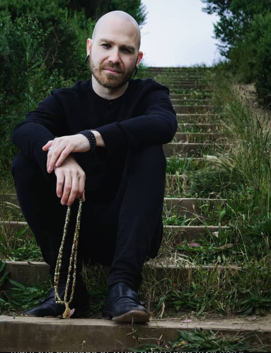 Shea Bilé wearing earrings sitting on outdoor stairs, dressed in black, holding a beaded necklace, with green bushes and trees in the background. San Francisco, CA.