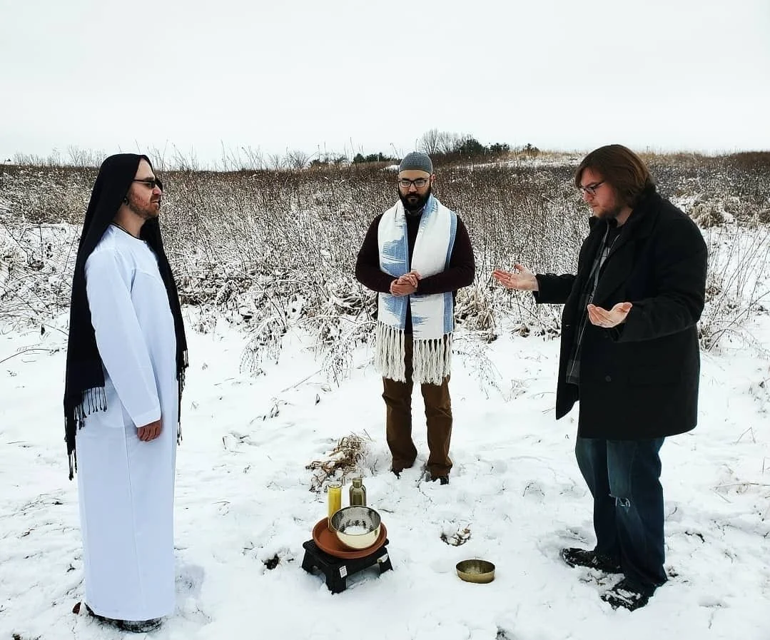 Shea Bilé, Samuel David, and another man wear religious attire participate in a ceremony outdoors on snow-covered ground, with ritual items including candles and bowls in front of them.