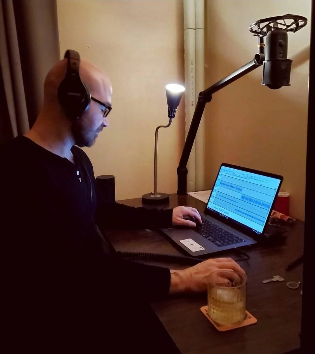 Shea Bilé wears headphones working on a laptop at a desk with a glass of drink, a small lamp, and a microphone arm setup.
