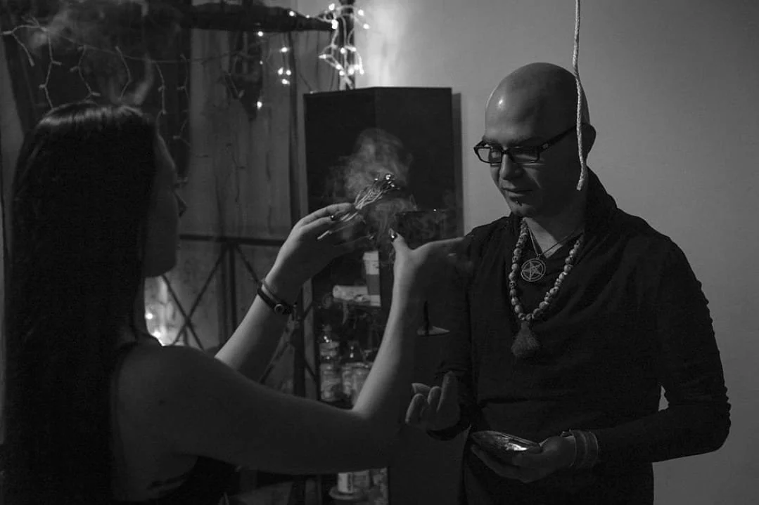 Luzia Lowe holds a lit incense stick in front of Shea Bilé, both standing indoors with fairy lights in the background. Shea wears layered necklaces, and a pentagram pendant.