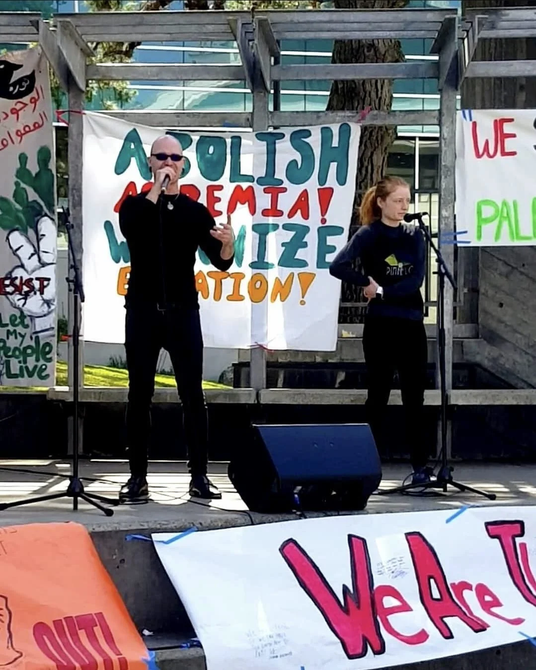 Shea Bilé stands on a stage with microphones, participating in a rally. Behind them are colorful banners with messages including "Stop War!" The setting is outdoors with trees and a building in the background. San Francisco, CA. SF State.