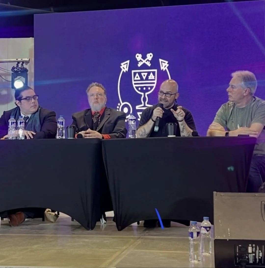 Shea Bilé and three other men sitting at a panel discussion, Shea Bilé is speaking into a microphone. There are water bottles on the table and a purple background with a graphic logo. TarotCon, Manila, Philippines.