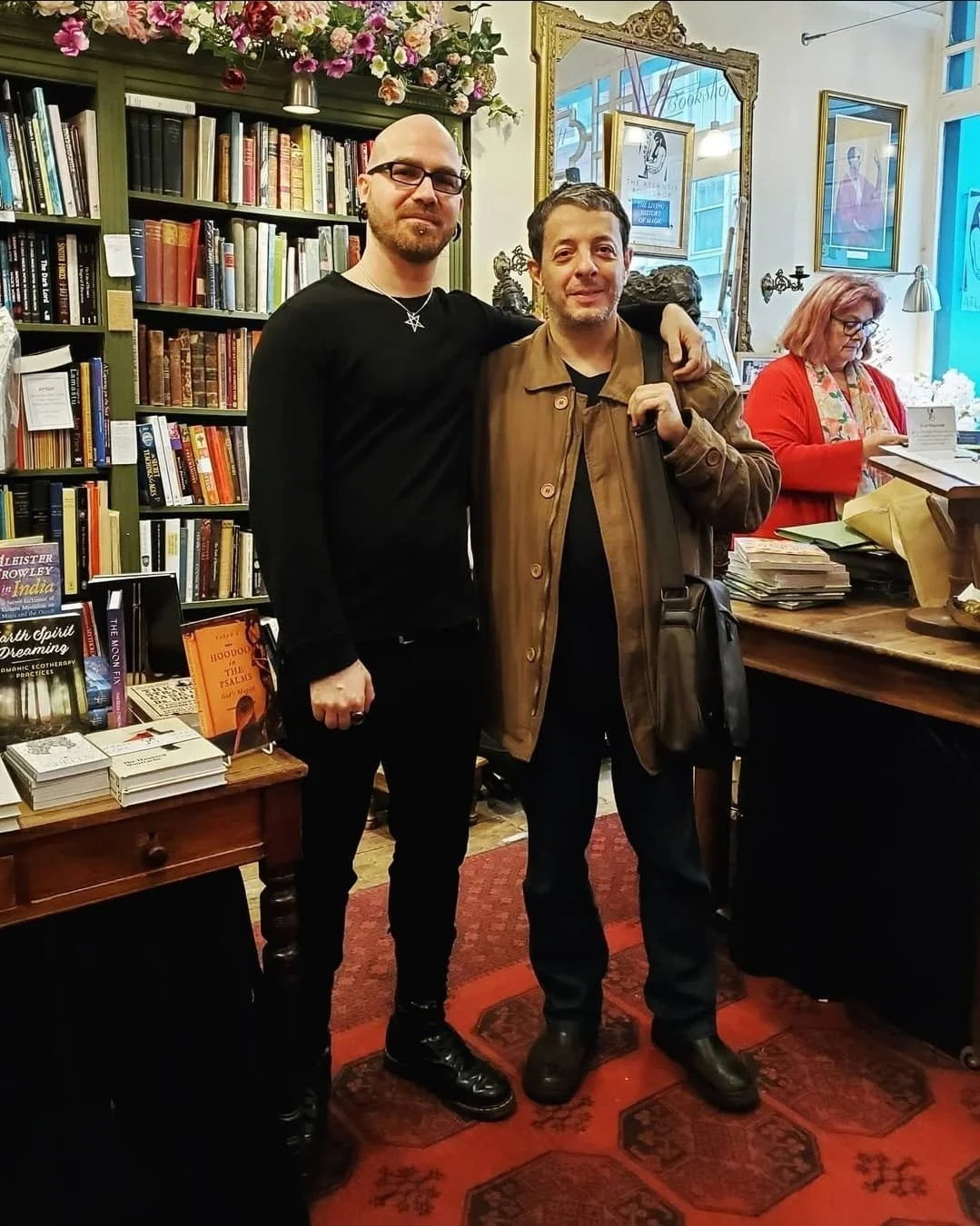 Shea Bilé and Humberto Maggi standing close together in the Atlantis bookstore in London, England, smiling, with bookshelves and a woman browsing in the background.