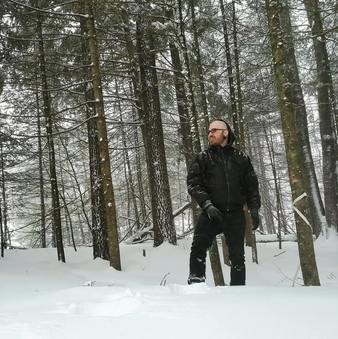 Shea Bilé dressed in black winter clothing stands in a snow-covered forest with tall trees, looking to the right. New Hampshire.