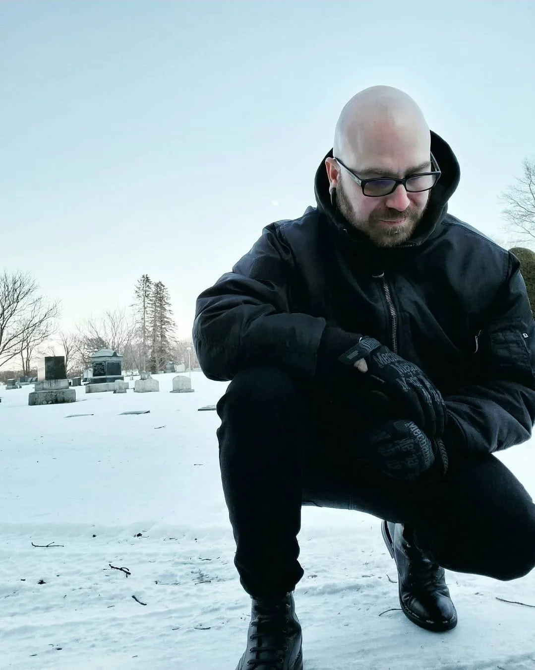 Shea Bilé is squatting on snow in a cemetery, wearing a black jacket, gloves, and boots, with gravestones and bare trees in the background.