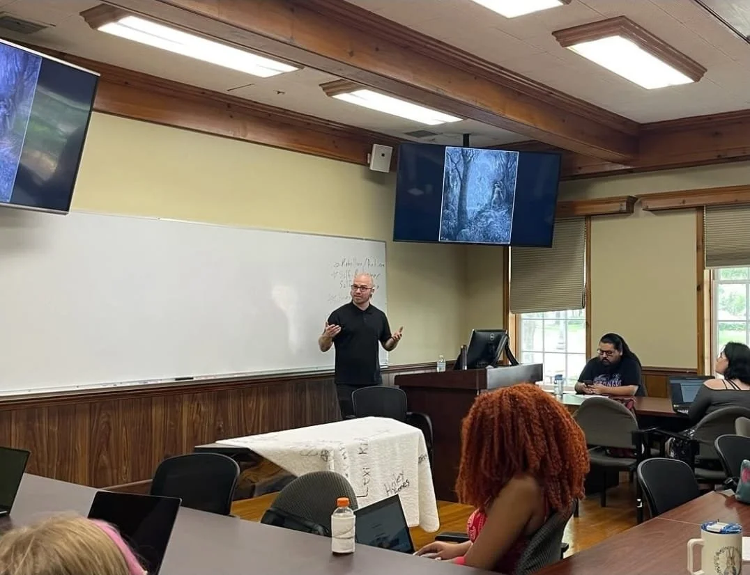 Shea Bilé gives a presentation at the front. There are several students are seated at tables. Two screens display an image of Lucifer in a forest. The room has wooden trim, beige walls, and large windows letting in natural light.