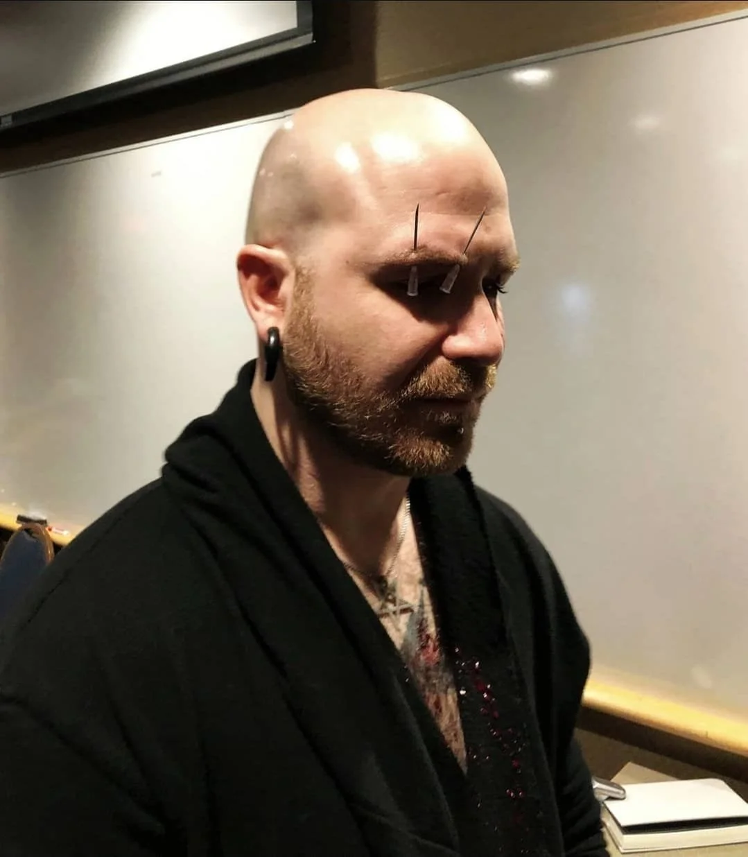 Shea Bilé stands with his eyes closed with needles inserted into his forehead, wearing black clothing and earrings, in an indoor setting. Paganicon, Minneapolis, MN.