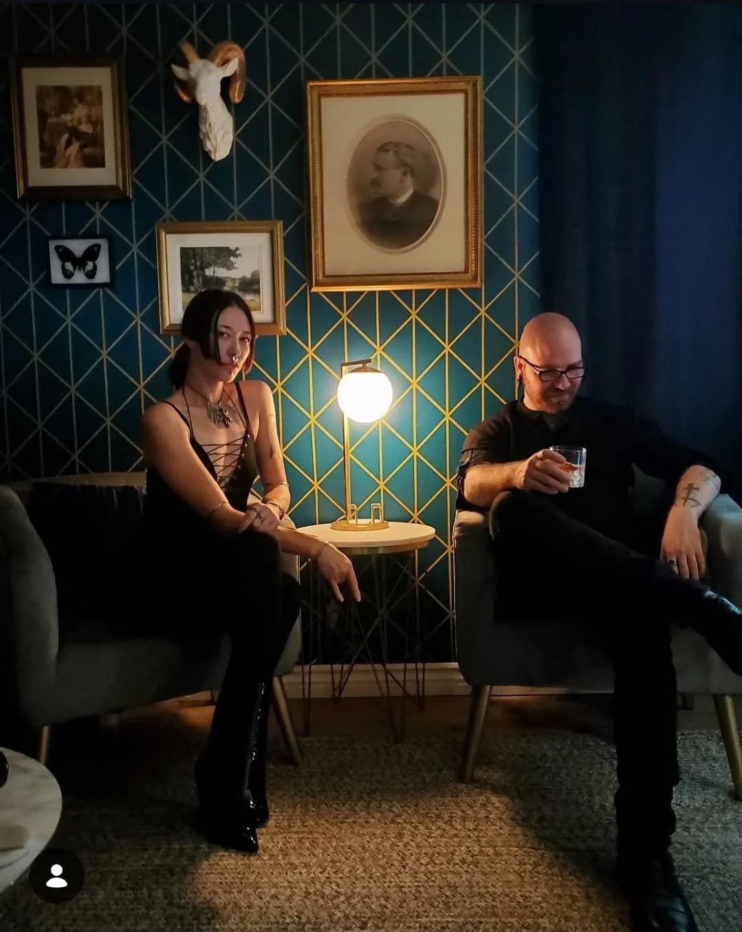 Shea Bilé and Luzia Lowe sitting on chairs in a dimly lit room, framed pictures and a mounted animal skull. Luzia is wearing a black outfit with a lace-up top, and Shea is wearing glasses and a black shirt, holding a glass.