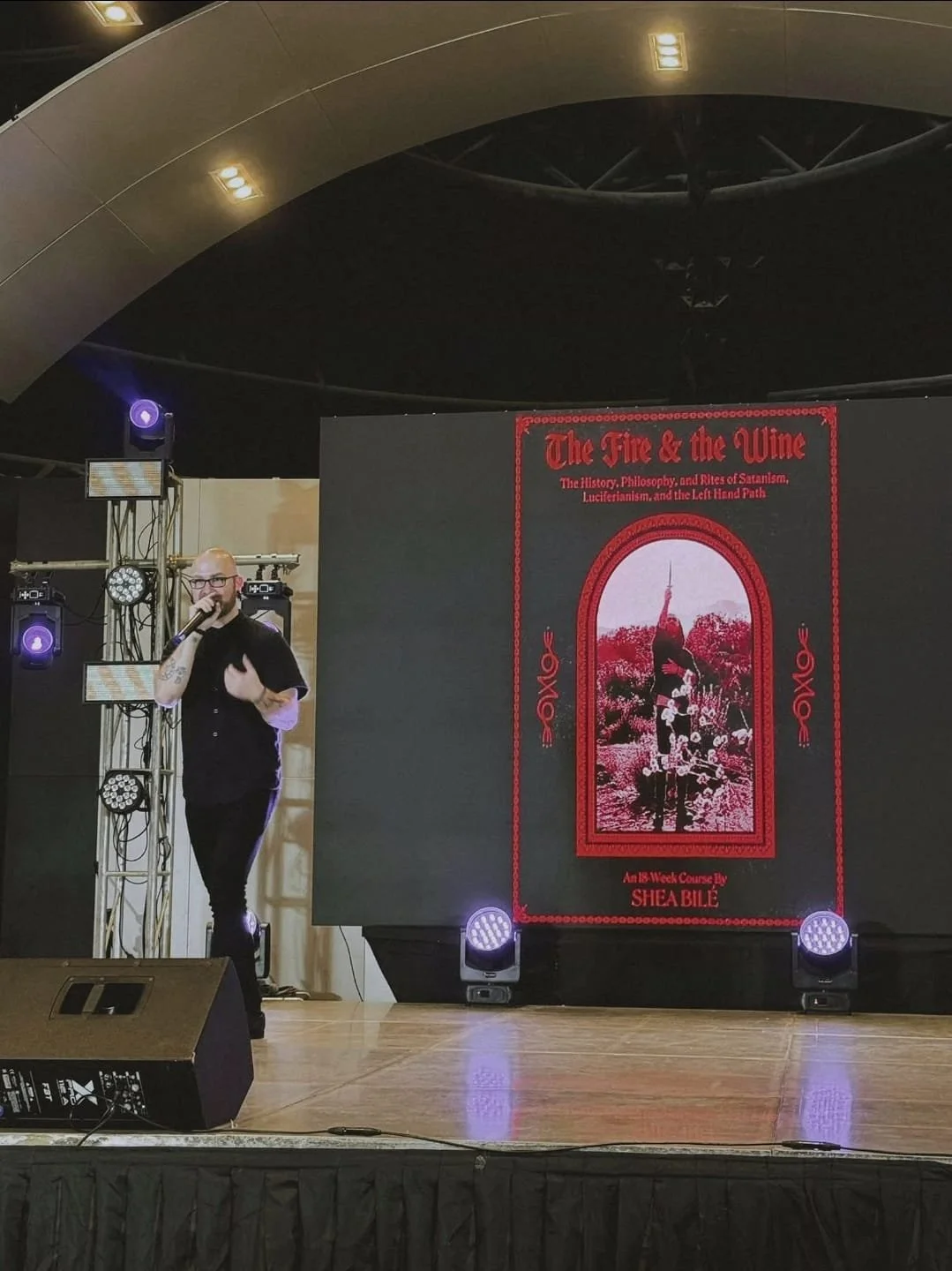 Shea Bilé is speaking into a microphone on a stage, with a large screen behind him displaying a promo image for 'The Fire & the Wine.' TarotCon. Manila, Philippines.