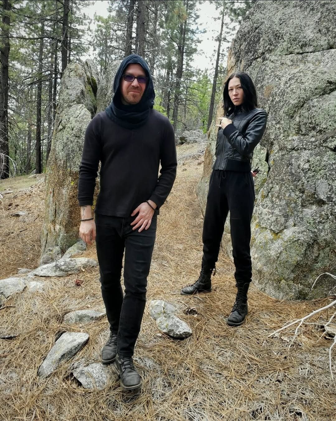 Shea Bilé and Luzia Lowe, standing in a forest among rocks and pine trees. Shea wears black clothing with a hood and sunglasses, and Luzia is in a black leather jacket and black pants, showing sign of horns. California.