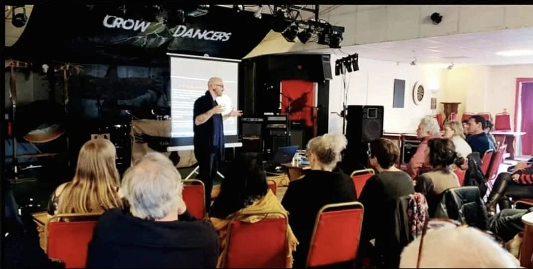 Shea Bilé presents at a crowd at Pandaemonium 2023 in Weymouth, England, standing in front of a projected screen, with an audience seated at tables.