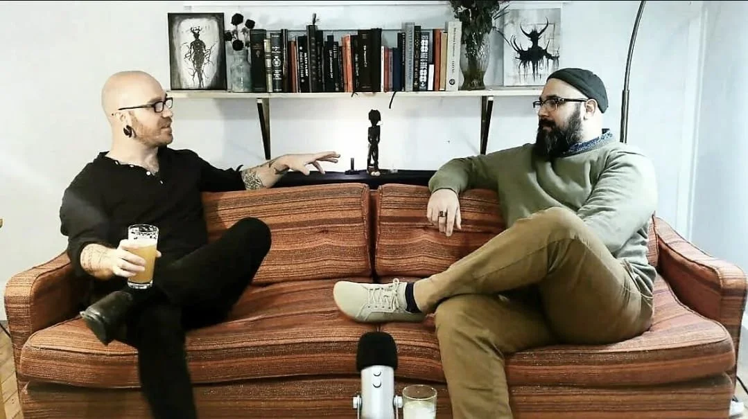 Shea Bilé and Samuel David on a vintage orange-brown striped sofa, engaged in a conversation. Shea is holding a beer. Samuel has a beard, glasses, a black beanie, and is dressed in a gray sweater. Behind them is a bookshelf with decorative items. 