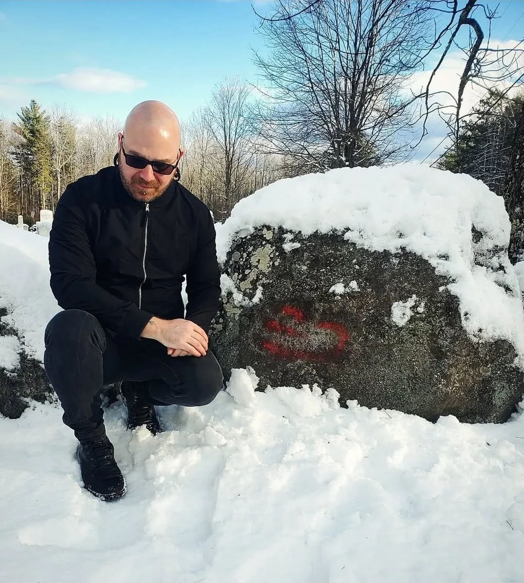 Shea Bilé is crouching next to a large snow-covered rock that has a red spray-painted Devil's Footprint on it, in a snowy outdoor setting with bare trees and a partly cloudy sky in the background. Augusta, Maine.