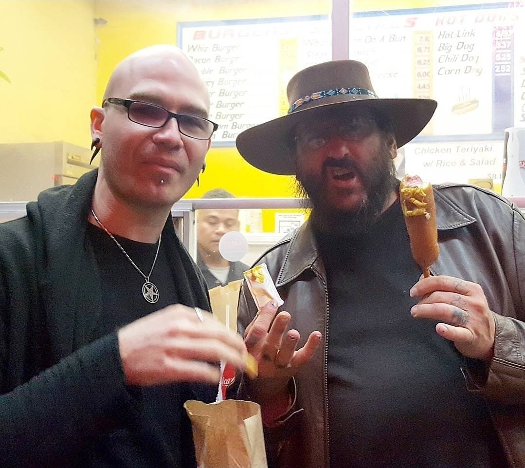 Shea Bilé and Steven Johnson Leyba stand outside a restaurant, Shea with glasses and a ring, Steven wearing a large hat and leather jacket, holding a corn dog, both holding food and looking at the camera. San Francisco, CA. Mission District. 