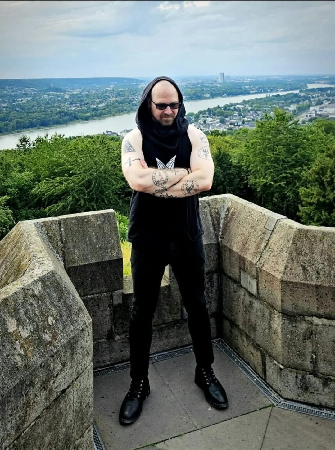 Shea Bilé on a stone platform, arms crossed, wearing black clothing, a sleeveless black shirt with a hood. The background features a scenic view of a river, green trees, and a cityscape with buildings. Cologne, Germany.