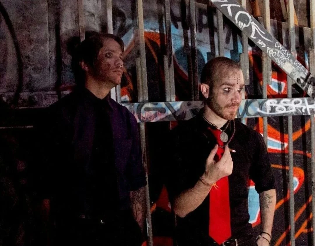 Shea Bilé and Nathan Gordon standing against a graffitied wall wearing dress clothing, with one adjusting his tie, and the other looking to the side.