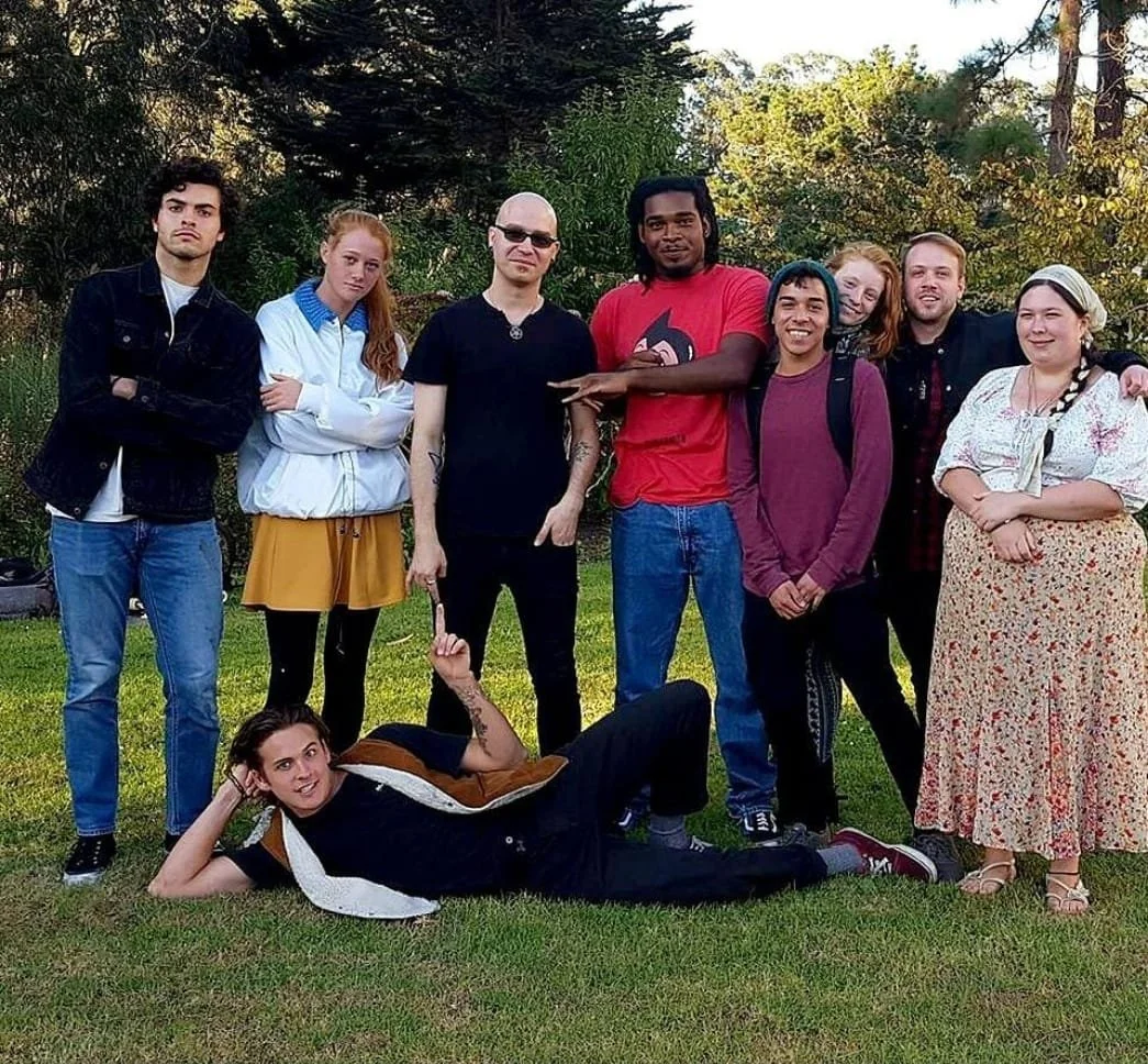 Shea Bilé and a group of ten diverse young adults posing outdoors on grass with trees in the background. One person is lying on the ground in front, while the others stand behind them. The Occult Student Alliance. San Francisco, CA.