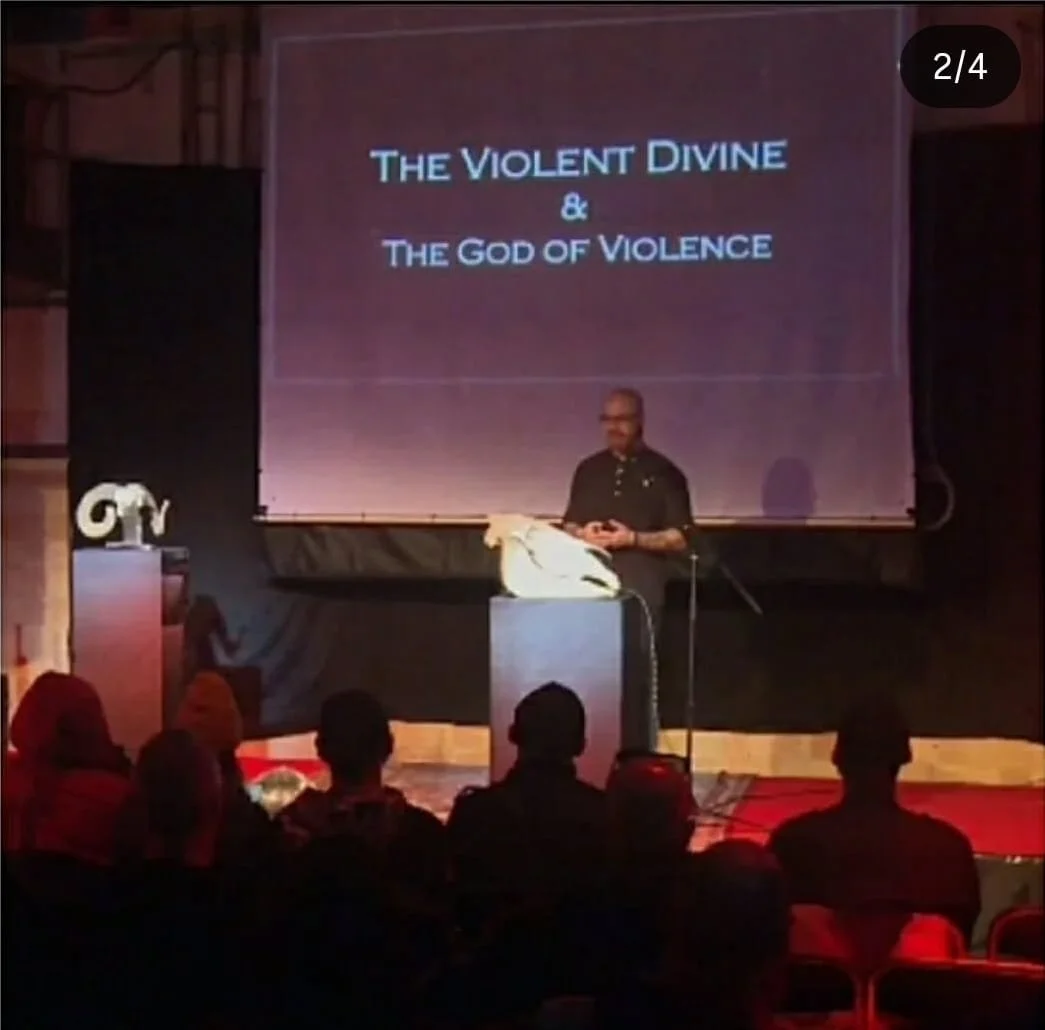 Shea Bilé gives a presentation titled "The Violent Divine & The God of Violence" on a stage in front of an audience. Occulture. Berlin, Germany.