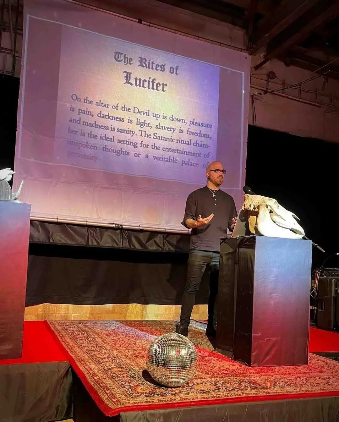Shea Bilé gives a presentation on a stage with a large screen behind him. The screen displays the title 'The Rites of Lucifer' and some text about Satanic rituals. The stage has a disco ball on the floor, and a podium with a goat skull. Occulture.