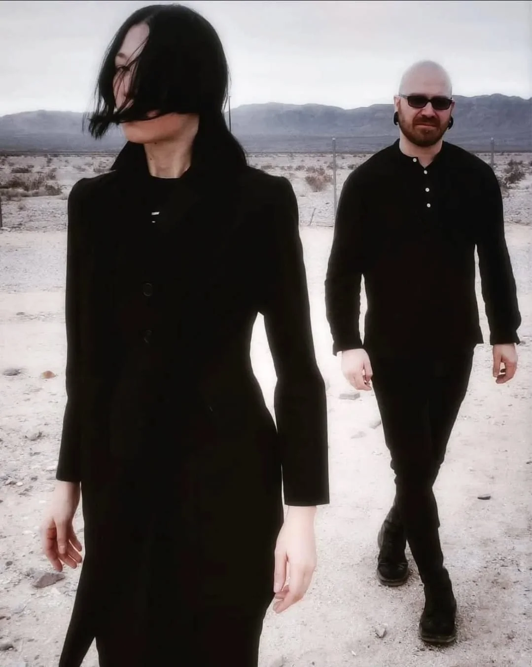 Shea Bilé and Luzia Lowe walking in a desert landscape with mountains in the background. The woman has black hair and is wearing a black dress, and the man has a beard, sunglasses, and is dressed in black. Mojave Desert. California. 