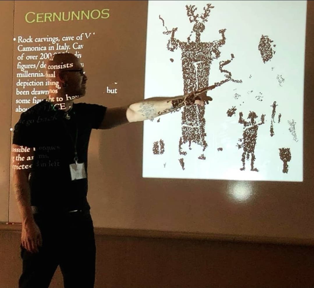 Shea Bilé is pointing at a projected image of rock carvings of a tree and human figures, with text about Cernunnos on a presentation slide. Paganicon.